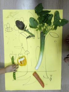 Disegno fatto da un bambino e sopra di esso varie verdure: carciofo, spinaci, porro, peperone, carota e piselli
