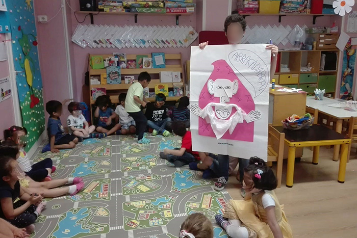 Bambini in cerchio che ascoltano la maestra, la quale mostra un cartellone con un mago disegnato che dice abracadabra
